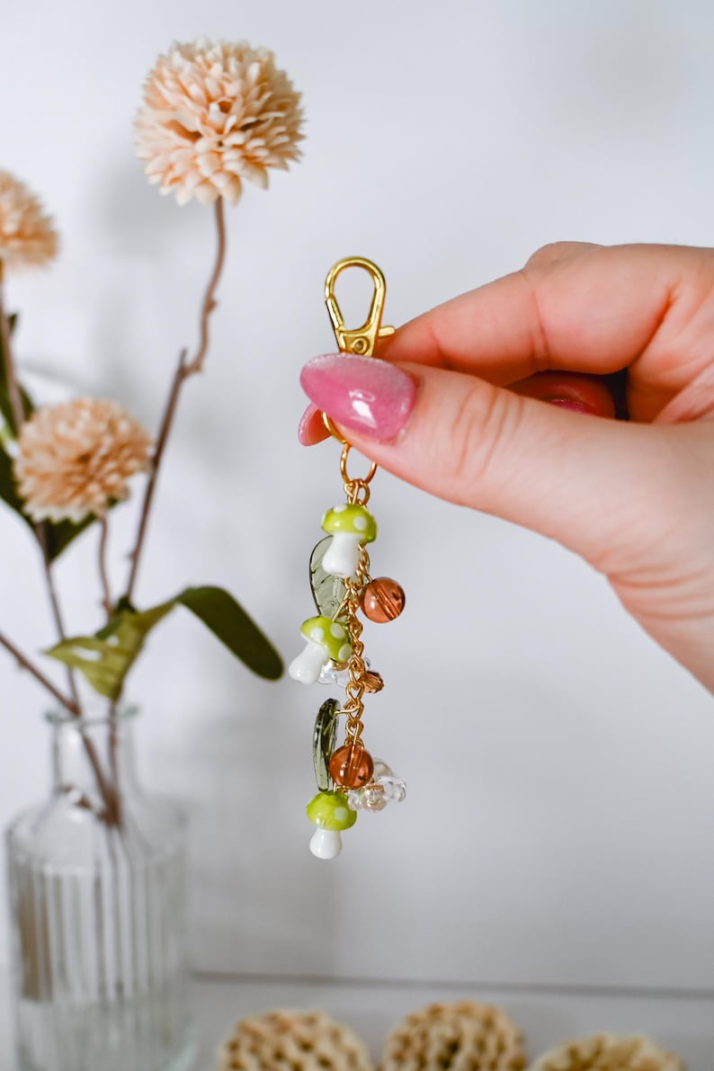 May include: A gold-colored keychain with a lobster clasp, adorned with mushroom charms in white and green, along with clear and amber-colored beads. The keychain is held in a hand, with a background of flowers in a vase.
