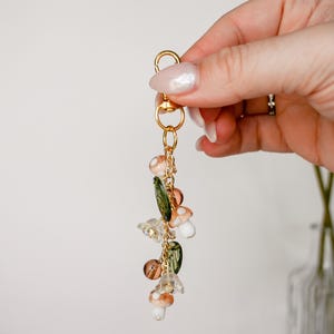 May include: A gold-colored keychain with a lobster clasp, adorned with mushroom and leaf charms. The charms are in shades of white, brown, and green, with clear and gold accents. The keychain is suspended from a gold chain.