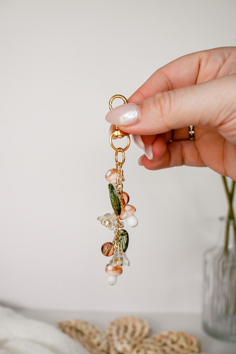 May include: A gold-colored keychain with a lobster clasp, adorned with mushroom and leaf charms. The charms are in shades of white, brown, and green, with clear and gold accents. The keychain is suspended from a gold chain.