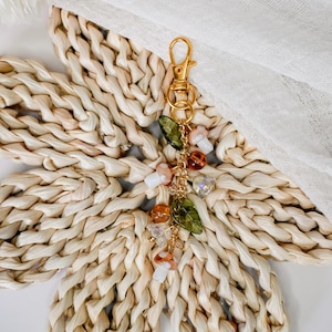 May include: A gold-colored keychain with a charm featuring mushroom and leaf-shaped beads. The charm includes clear, orange, and white beads, and green leaf-shaped beads. The keychain is displayed on a woven, flower-shaped mat.