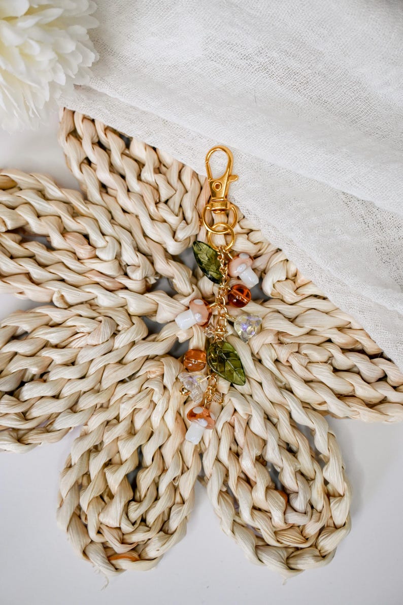 May include: A gold-colored keychain with a charm featuring mushroom and leaf-shaped beads. The charm includes clear, orange, and white beads, and green leaf-shaped beads. The keychain is displayed on a woven, flower-shaped mat.