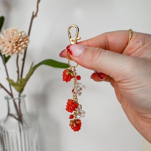 Puede incluir: Un llavero dorado con cierre de mosquetón, adornado con frambuesas de cuentas rojas y cuentas de cristal transparente. El llavero está suspendido de una cadena dorada. Un accesorio encantador.