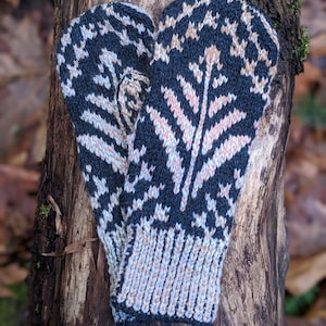 May include: A pair of knitted mittens with a dark navy blue background and a floral pattern in light pink, beige, and white. The cuffs are ribbed in a light blue and beige color. The mittens are displayed on a tree trunk.