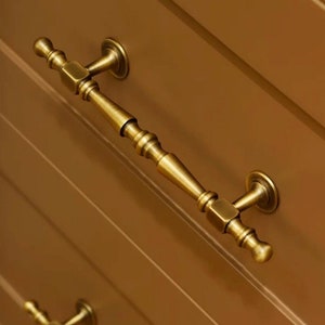 May include: Two antique brass cabinet drawer pulls with a decorative design. The pulls are mounted on a wooden cabinet door.