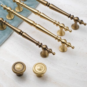 May include: Four gold-toned cabinet pulls with ornate detailing and two matching knobs. The pulls are all different lengths and have a slightly aged finish.