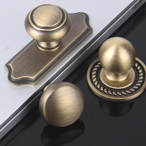 May include: Close-up of three antique brass door knobs. One knob is mounted on a rectangular backplate, while the other two are round. The knobs have a vintage, aged appearance, with a warm, metallic finish. The design is classic and elegant.