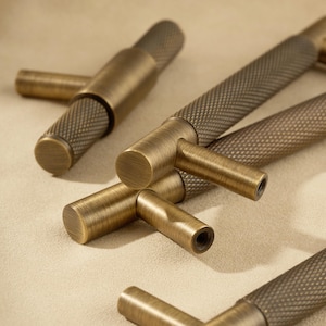 May include: Close-up of several antique brass cabinet pulls. The pulls have a textured, diamond-patterned grip and a cylindrical handle. The hardware is a warm, muted gold colour, suggesting a vintage aesthetic. The pulls are arranged on a neutral background.