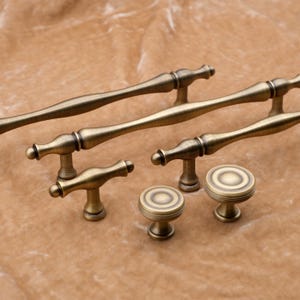 May include: A set of antique brass cabinet hardware, including two long pulls, two smaller pulls, and two knobs. The hardware has a distressed finish and a classic design.
