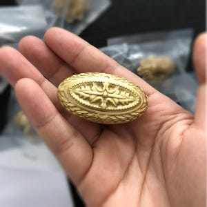 K&ouml;nnte beinhalten: Ein goldfarbener, ovaler Dekorknopf mit einem komplizierten floralen Design.