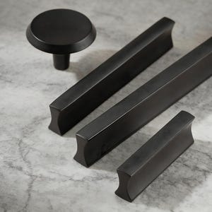 May include: A collection of matte black cabinet hardware, including a round knob and three rectangular bar pulls of varying lengths. The hardware is displayed on a light gray surface, showcasing a modern design aesthetic.