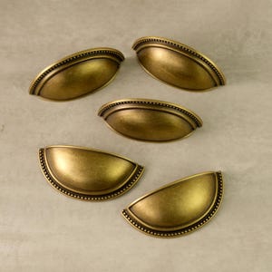 Könnte beinhalten: Fünf antike Schubladengriffe in Messingoptik auf einer neutralen Oberfläche. Jeder Griff hat eine halbmondförmige Form mit einem Perlenrand und einer gewölbten, vertieften Mitte. Diese Griffe sind für Möbel und Schränke konzipiert.