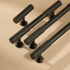 May include: A set of four black cabinet pulls with a ribbed design. The pulls have a cylindrical shape with a rounded base and are made of metal. The pulls are arranged on a neutral background, showcasing their modern design.