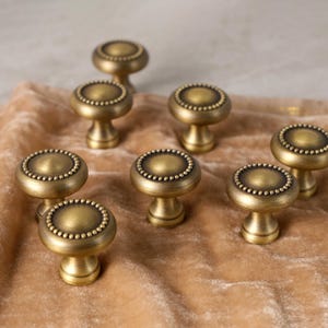 May include: Seven antique brass cabinet knobs are displayed. Each knob has a round, beaded edge and a short, pedestal base. They are arranged on a tan velvet surface, giving a vintage feel. The knobs are made of brass.