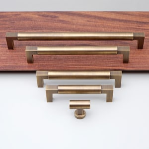 Puede incluir: Cinco tiradores de armario de latón antiguo en varios tamaños, mostrados en una superficie de madera. Los tiradores tienen una base cuadrada y un mango cilíndrico.