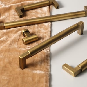 Può includere: Primo piano di diverse maniglie per mobili e un pomello in ottone antico. L'hardware ha un design rettangolare con finitura spazzolata. Le maniglie sono di diverse lunghezze e il pomello ha una forma piccola e rotonda. L'hardware è esposto su una superficie di velluto beige.