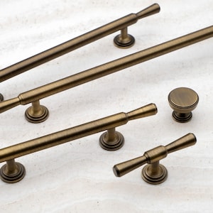 May include: Assortment of antique brass cabinet hardware, including pulls and knobs. The pulls have a simple, elegant design with a tapered end. The knobs are round with a raised edge.