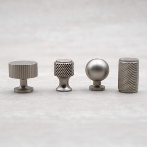 May include: Four different metal cabinet knobs in a brushed nickel finish. The knobs are in a row from left to right: a round knob with a ridged design, a round knob with a diamond pattern, a round knob with a smooth surface, and a cylindrical knob with a diamond pattern.