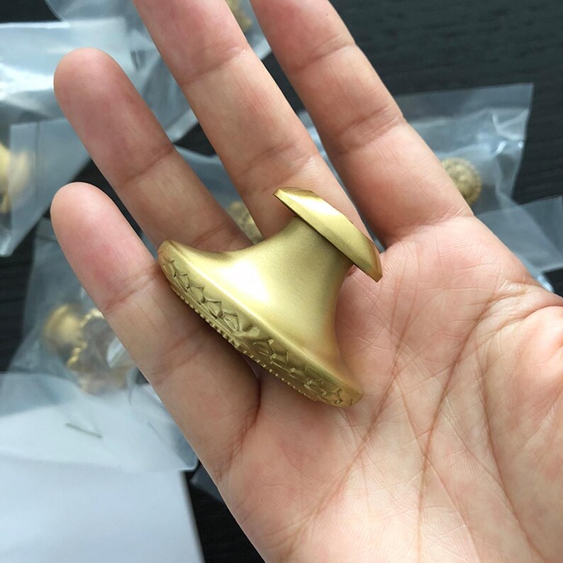 K&ouml;nnte beinhalten: Ein goldfarbener Schrank-Knopf mit einem dekorativen, verzierten Design um die Basis. Der Knopf hat eine abgerundete Oberseite und eine leicht erh&ouml;hte Mitte.
