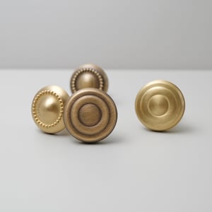 Könnte beinhalten: Vier antike Messing-Schrankknöpfe in verschiedenen Designs. Ein Knopf hat einen Perlenrand, ein anderer konzentrische Kreise, und zwei haben ein glattes, abgerundetes Design. Die Knöpfe sind in einem warmen, metallischen Finish.