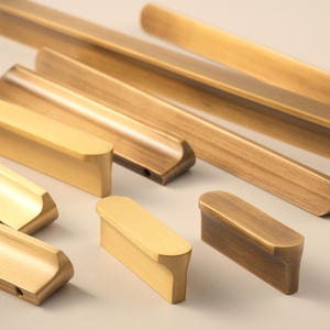 Op de afbeelding: Een verzameling messing kastknoppen en handgrepen in verschillende maten en vormen. De hardware heeft een warme, gouden tint en een geborstelde afwerking. Sommige knoppen zijn rechthoekig, andere lang en gebogen.