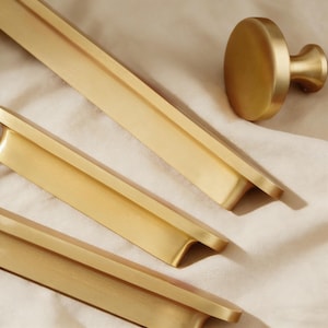 May include: Close-up of several gold-colored cabinet pulls and a round knob. The hardware has a brushed finish and a modern design. The pulls are rectangular with a curved edge. The knob is circular with a short, cylindrical stem.