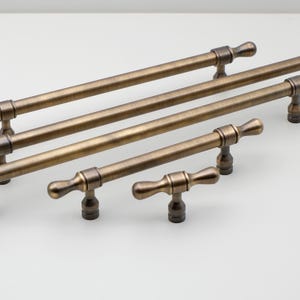 May include: Five antique brass cabinet pulls with a t-shaped design. The pulls have a distressed finish and are available in various lengths.