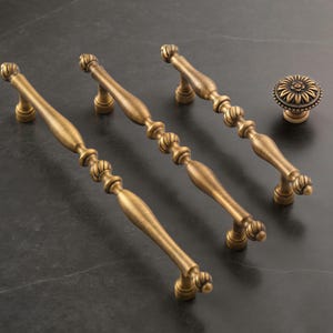 May include: Four antique brass cabinet pulls and a matching knob. The pulls have a decorative, elongated design with a series of rounded accents. The knob features a floral pattern. All pieces have a warm, aged finish.