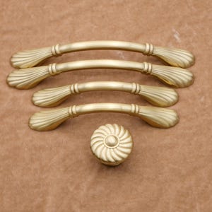 Könnte beinhalten: Satz mit vier goldenen Schrankgriffen mit einem dekorativen Muschel-Design und einem passenden goldenen Knopf. Die Griffe sind gebogen und haben eine glatte Oberfläche.