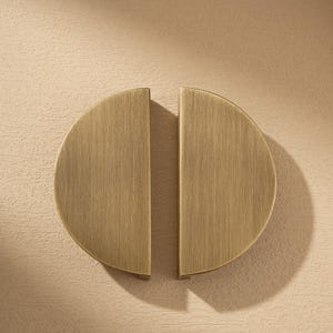 May include: Two brass-colored, semi-circular cabinet pulls. Each pull has a brushed metal finish and is mounted on a light beige textured surface. The pulls are arranged to form a complete circle.