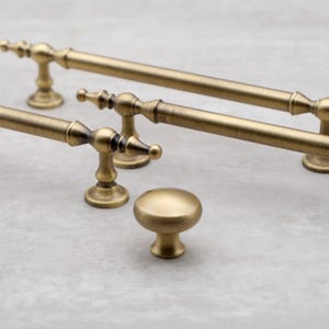 May include: Close-up of antique brass cabinet hardware, including two bar pulls and a round knob. The hardware has a vintage aesthetic with ornate detailing and a warm, golden hue. The pulls are approximately 8 inches long.