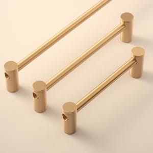 May include: Three gold-colored cabinet pulls with a cylindrical design. Each pull has a straight bar connecting two rounded cylinders, with a small hole on one cylinder. The pulls are arranged diagonally against a neutral background.