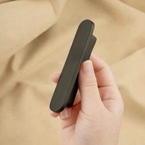 May include: A sleek, matte black cabinet pull with a rounded end and a small, curved handle. The pull is held in a hand against a beige background. This modern hardware is suitable for drawers and cabinets.