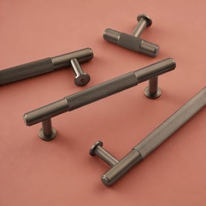 May include: A collection of gunmetal cabinet pulls with a ribbed design, set against a pink backdrop. The pulls feature a cylindrical body and a T-shaped handle. These decorative hardware pieces are for home improvement.