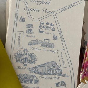 Puede incluir: Una tarjeta de color crema con un mapa en tinta azul del lugar Deerfield Estates Venue. El mapa ilustra carreteras, Cabins 1-6, Cabins 7&8, un área de ceremonia al aire libre, una capilla, un salón de recepción y otros edificios etiquetados.