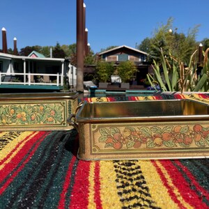May include: Two rectangular metal containers with gold trim and handles. The sides feature a decorative design of green leaves and orange fruit. They sit on a colourful striped blanket, with a waterfront scene in the background.