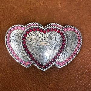May include: Silver-toned heart-shaped belt buckle with pink and red gemstones. The buckle features intricate floral scrollwork and a rope-like border. The three hearts are joined together, creating a unique design.