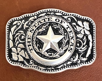 May include: A rectangular silver-toned belt buckle with scalloped edges featuring a central five-pointed star surrounded by a circular border with the text THE STATE OF TEXAS. The background is black with embossed floral and scrollwork patterns.