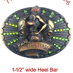May include: A bronze-colored belt buckle with a 1 1/2 inch wide heel bar. The buckle features a detailed design of a woman wearing a gas mask and holding a skull. The buckle is decorated with green rhinestones and the text "The Devil Comes Smiling".