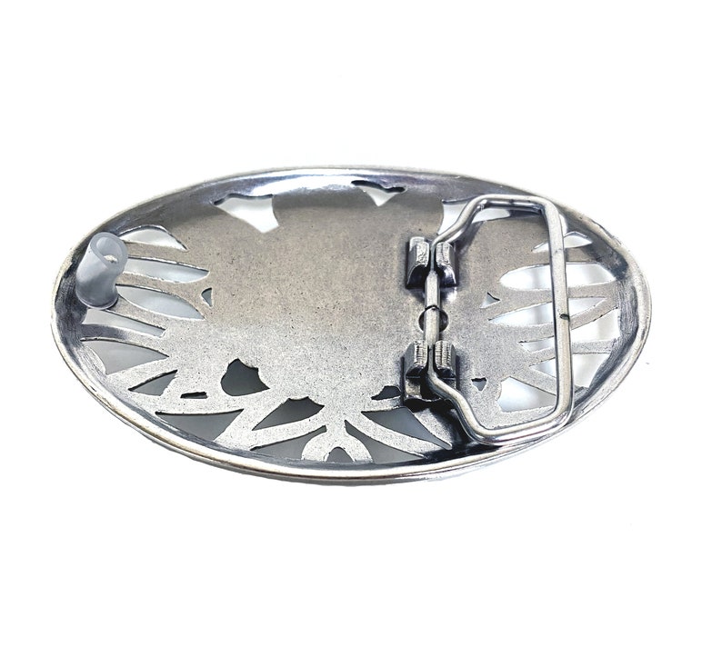 May include: Silver-toned metal belt buckle with an oval shape and a floral design. The buckle has a metal loop for attaching a belt.