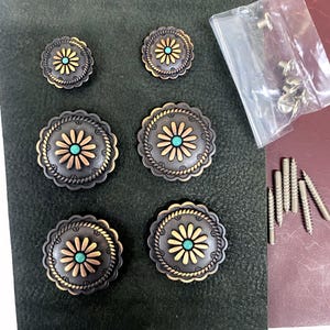 May include: A collection of decorative metal conchos with a floral design. The conchos have a dark gray base with gold-colored accents and a turquoise center. A small bag of screws and several long screws are also present.