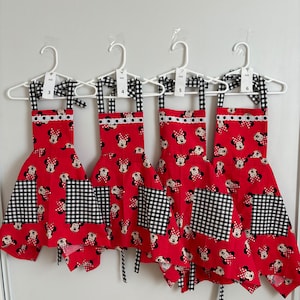 May include: Four red aprons with a Minnie Mouse pattern, black and white gingham accents, and white lace trim. Each apron is labeled with a size number from 3 to 6 and hangs on a white hanger. The aprons have black and white gingham bows.