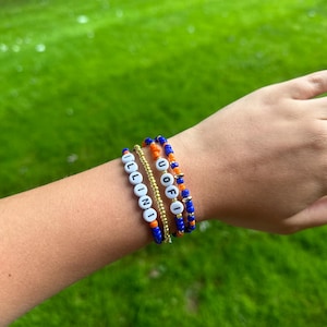 May include: Four beaded bracelets in blue, orange, and gold. One bracelet spells "ILLINI" in white beads. Another spells "U OF I". The bracelets are stacked on a wrist against a green background.