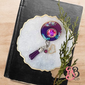 May include: A purple and pink resin charm with glitter and a heart charm attached to a purple tassel. The charm is on a white background with a gold rim.