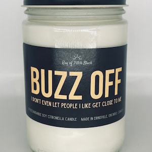 May include: A clear glass jar candle with a black lid and a black label. The label reads "BUZZ OFF" in gold lettering, with the text "I don't even let people I like get close to me" below. The candle is filled with white wax.