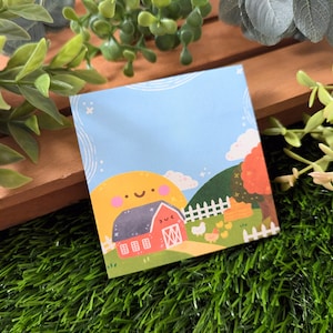 May include: A blue notepad with a cute cartoon illustration of a red barn with a yellow sun and white fence. The barn has a smiling face and the sun has rosy cheeks. The notepad is surrounded by green grass and a small tree with orange leaves.