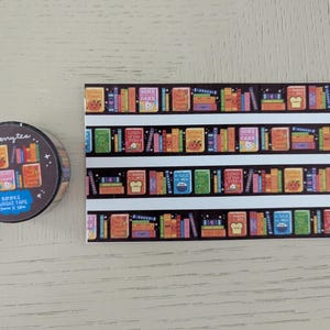 K&ouml;nnte beinhalten: Eine Rolle Washi-Tape und ein rechteckiges Blatt mit einem B&uuml;cherregal-Design. Die Klebebandrolle ist dunkelbraun mit einem sich wiederholenden Muster von B&uuml;chern. Das Blatt zeigt einen schwarzen Hintergrund mit wei&szlig;en Regalen und bunten B&uuml;chern.