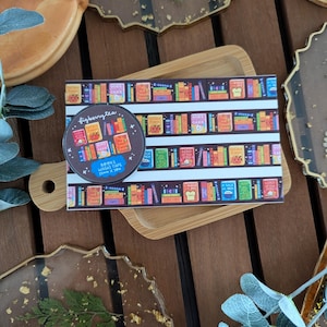 K&ouml;nnte beinhalten: Eine Rolle Washi-Tape und ein Aufkleber mit einem B&uuml;cherregal-Design mit dem Text "figberrytea". Das Klebeband ist 15 mm x 10 m gro&szlig;. Die Artikel befinden sich auf einem Holztablett und sind von dekorativen Harzuntersetzern umgeben.