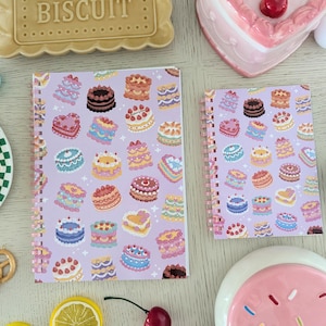 May include: Two spiral-bound notebooks with a lavender background and a pattern of colorful cakes. The notebooks are surrounded by decorative items, including a biscuit, a plate, and a heart-shaped dish.