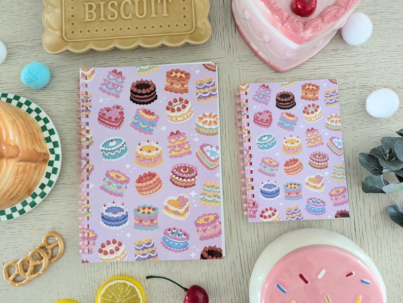 May include: Two spiral-bound notebooks with a lavender background and a pattern of colorful cakes. The notebooks are surrounded by decorative items, including a biscuit, a plate, and a heart-shaped dish.
