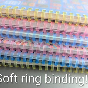May include: A stack of notebooks with soft ring binding. The notebooks have clear plastic covers and colourful spiral bindings in yellow, pink, and blue. The text "Soft ring binding!!" is visible at the bottom of the image.